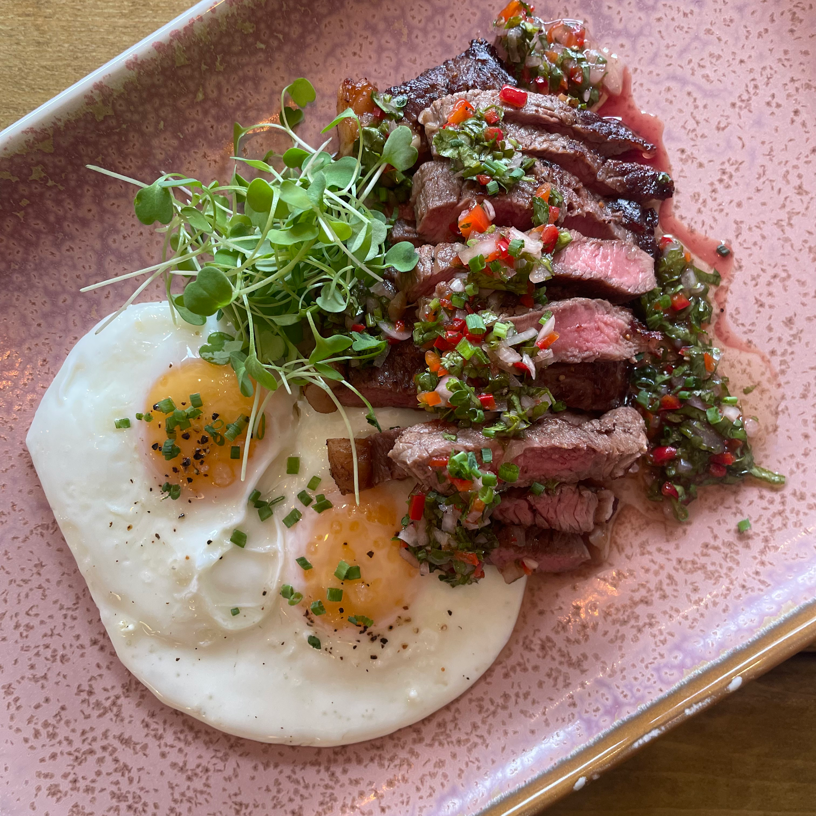 Sliced steak with herb salsa and two fried eggs with microgreens on a pink speckled plate.