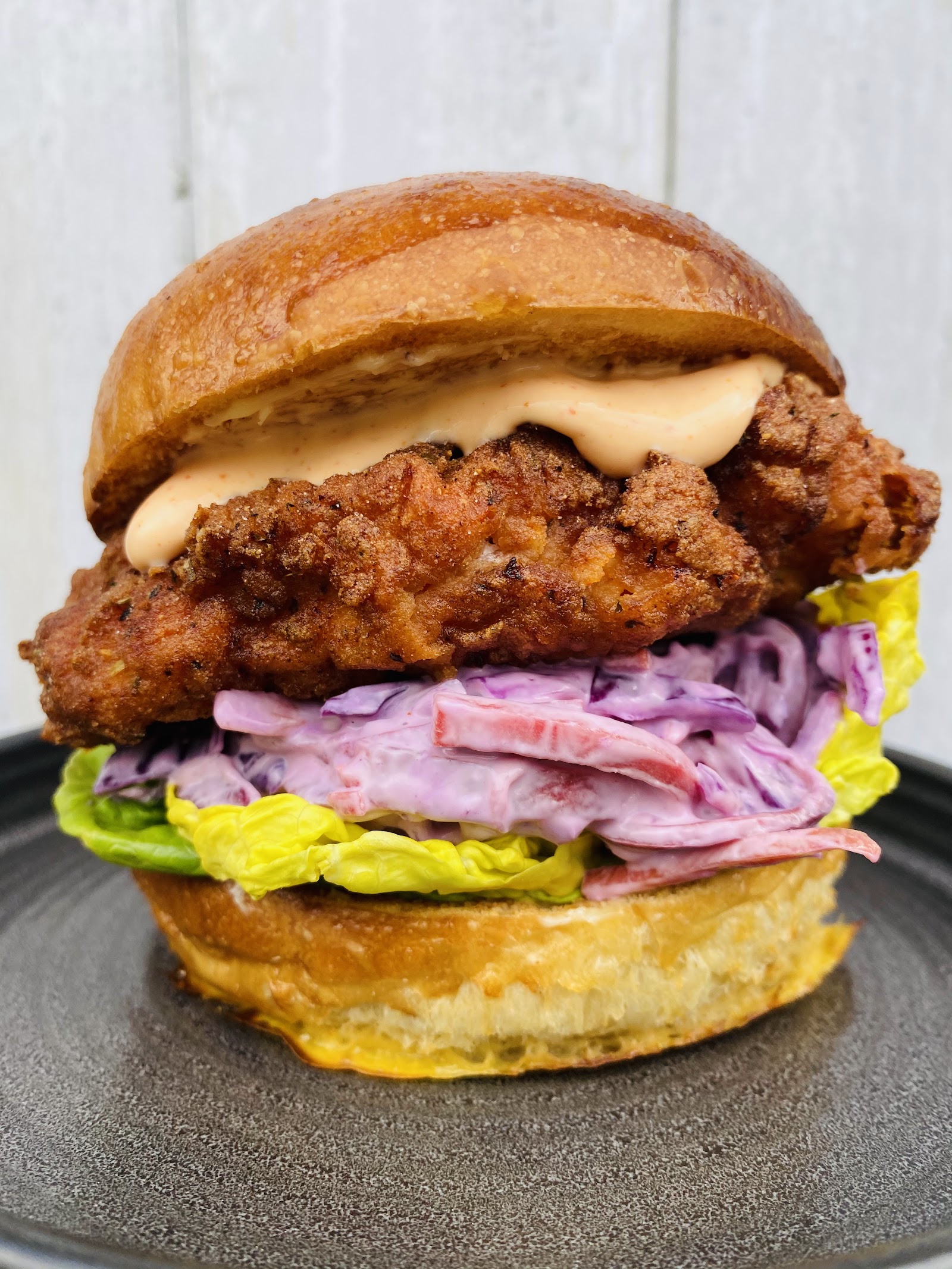 A tall fried chicken brioche burger with slaw and sauce on a dark plate.